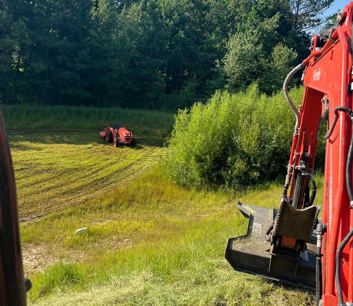 Excavator and tractor working together to grade a sloped area near a wooded landscape